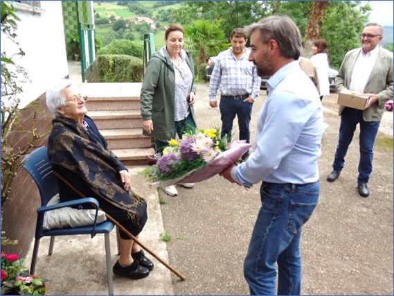 En el momento de recibir del Alcalde el ramo de flores, con Humberto Vigil esperando su turno para entregarle un presente de la Parroquia Rural. Al fondo, su nieto, Javier Ramos, y la esposa de ste, Olga Arboleya. | EL ECO